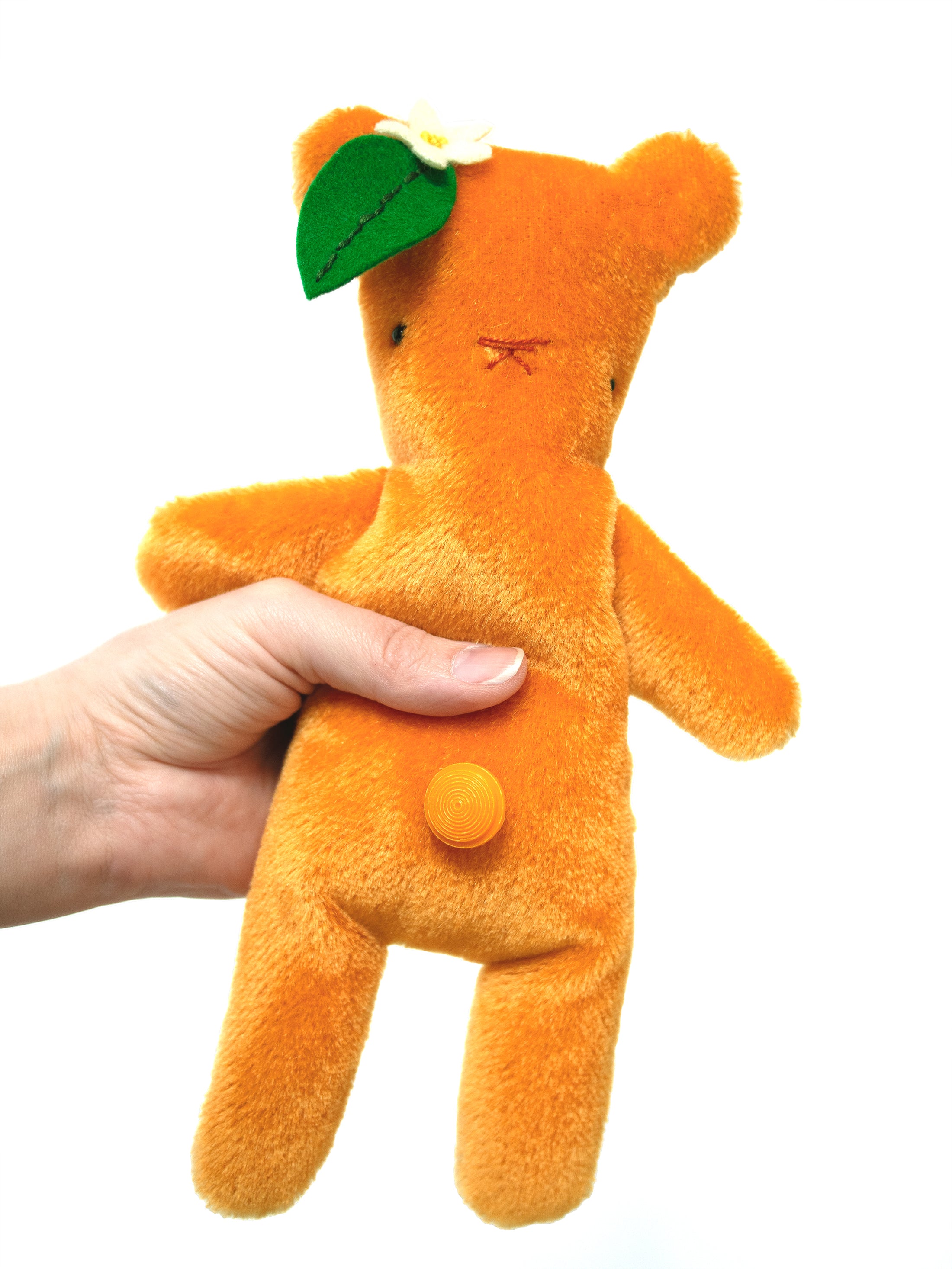 This is Orange by Bear Bears. The plush is covered in short, dense, slightly glossy fur that lies smooth with no visible curl; the coat colour is orange overall, including the muzzle, inner ears, tummy and paws which match the coat. The face has two small black plastic bead eyes set close to the centre and an orange-brown stitched X-shaped mouth/nostril detail rather than a separate moulded nose. A green felt leaf with visible darker stitching and a small white felt flower with a yellow centre are atta...